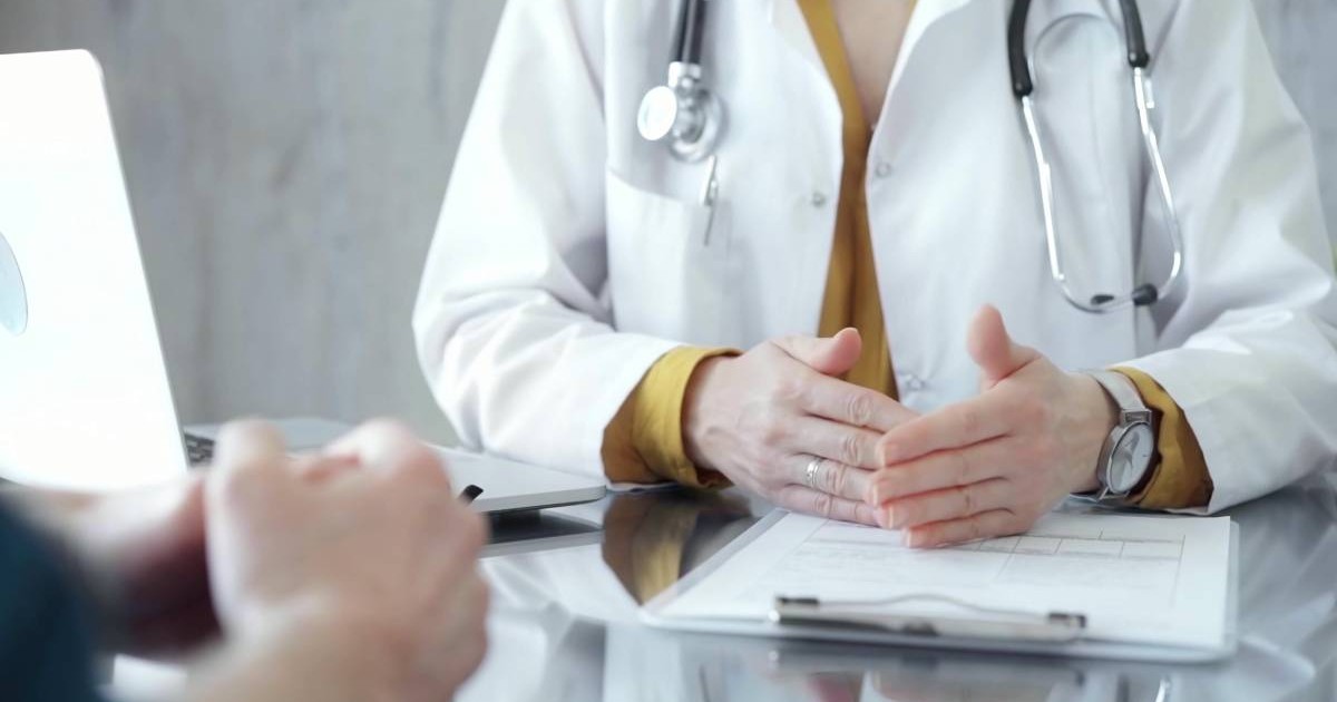 Medic îmbrăcat în halat alb, purtând un stetoscop în jurul gâtului și un ceas de mână, gesticulând ușor deasupra unei mese pe care se află o fișă medicală și un laptop deschis.