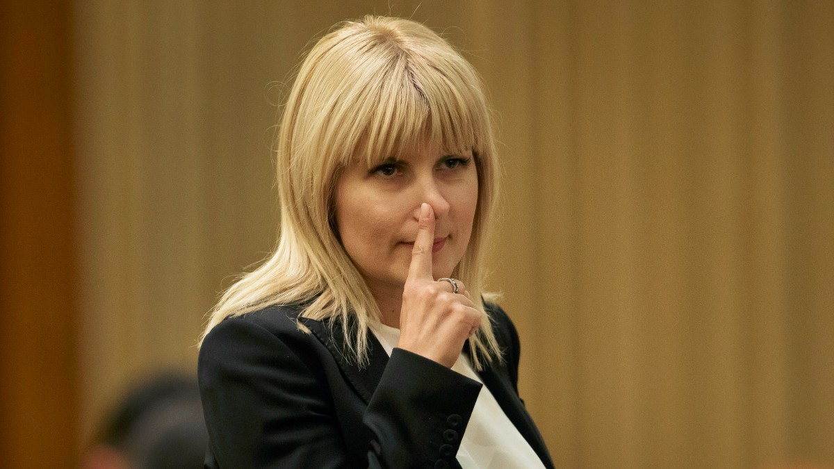 Imaginea o surprinde pe Elena Udrea într-un cadru oficial, îmbrăcată într-un sacou negru și o bluză albă, în timp ce își duce degetul la nas într-un gest care sugerează reflecție sau concentrare. Fotografia pare realizată în timpul unei ședințe sau al unui eveniment public, având un fundal discret și o atmosferă sobră.