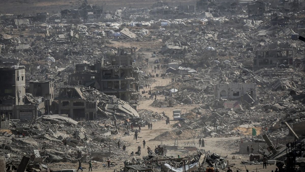 Fotografia realizată în Fâșia Gaza pe data de 16 octombrie 2025, după retragerea armatei israeliene din orașul Gaza conform acordului de încetare a focului, prezintă o zonă urbană distrusă aproape complet. Se văd clădiri prăbușite, mormane de ruine și structuri grav avariate, întinse pe o suprafață vastă. Printre ruine, pe o arteră centrală acoperită de praf, se observă siluetele unui număr mare de persoane, alături de câteva vehicule albe