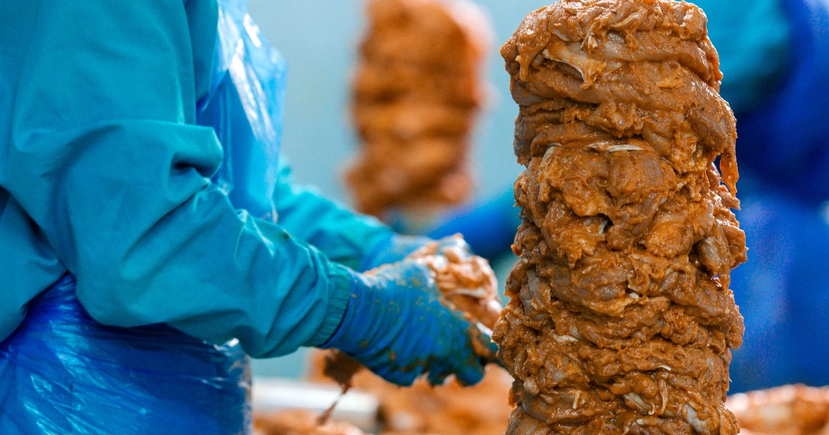 Imaginea surprinde procesul de preparare a cărnii pentru kebab într-o unitate de producție alimentară, unde angajații lucrează în condiții controlate de igienă. În prim-plan se observă un rotisor de carne destinat produselor de tip kebab, pregătit pentru distribuție către restaurante și comercianți.