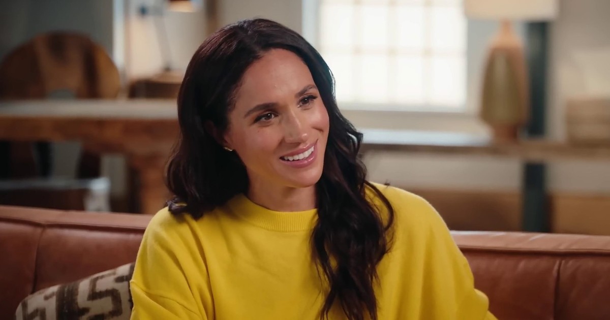 Meghan Markle, zâmbind, îmbrăcată într-un tricou galben, cu capul ușor într-o parte