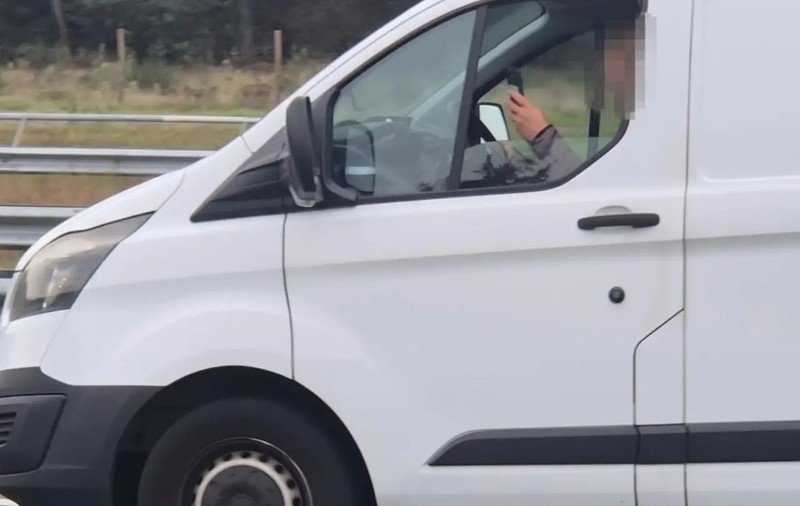 Imaginea prezintă un șofer (cu fața blurată) la volanul unei autoutilitare albe, utilizând un telefon mobil în timp ce conduce pe autostradă.