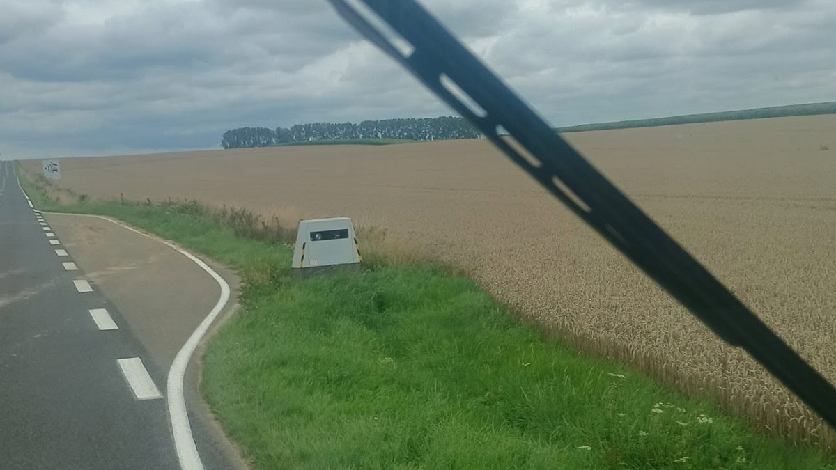 Un radar autonom amplasat pe marginea unui drum rural, lângă un câmp, văzut din interiorul unui vehicul prin parbriz.   