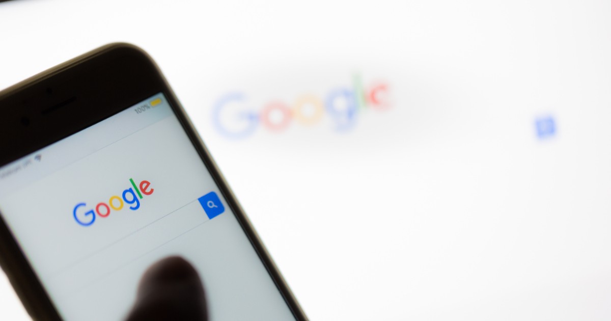 Un telefon ținut în mână, aflat în prim-plan, pe ecranul căruia este deschisă pagina principală Google. În fundal apare un al doilea ecran care afișează logo-ul Google.