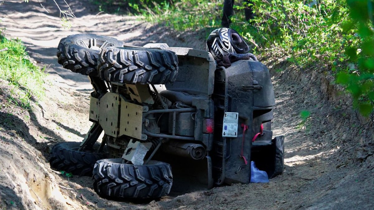 Un ATV negru răsturnat pe partea dreaptă pe un drum din pădure, cu iarbă verde în jur