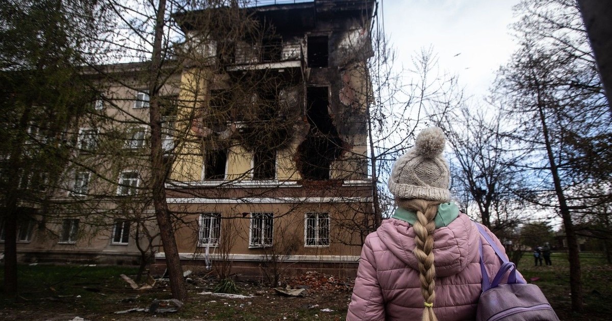 Femeie cu spatele, îmbrăcată cu geacă roz, cu fes pe cap, care se uită la o clădirre distrusă de bombardamente rusești