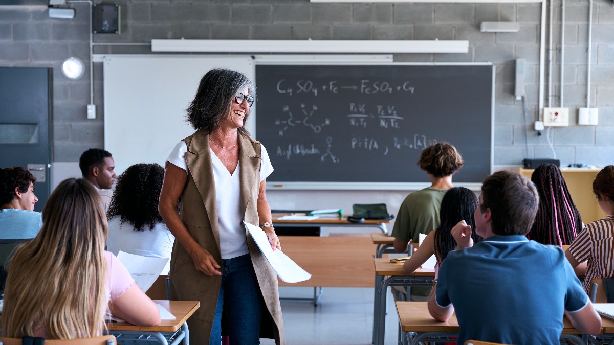 O profesoară zâmbitoare interacționează cu elevii într-o sală de clasă luminoasă, creând o atmosferă relaxată și deschisă. Imaginea surprinde un moment de implicare activă, în care elevii par atenți și interesați de desfășurarea orei.