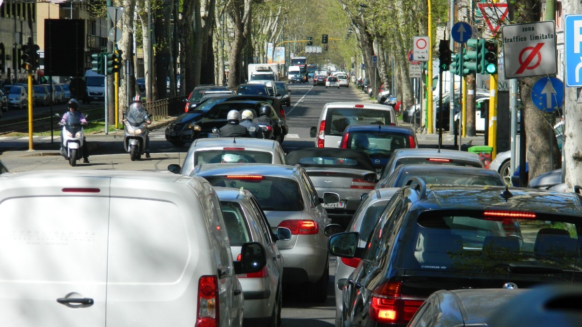 Trafic aglomerat pe un bulevard urban mărginit de copaci, cu numeroase mașini și scutere oprite sau deplasându-se în coloană lângă un semafor și indicatoare rutiere.
