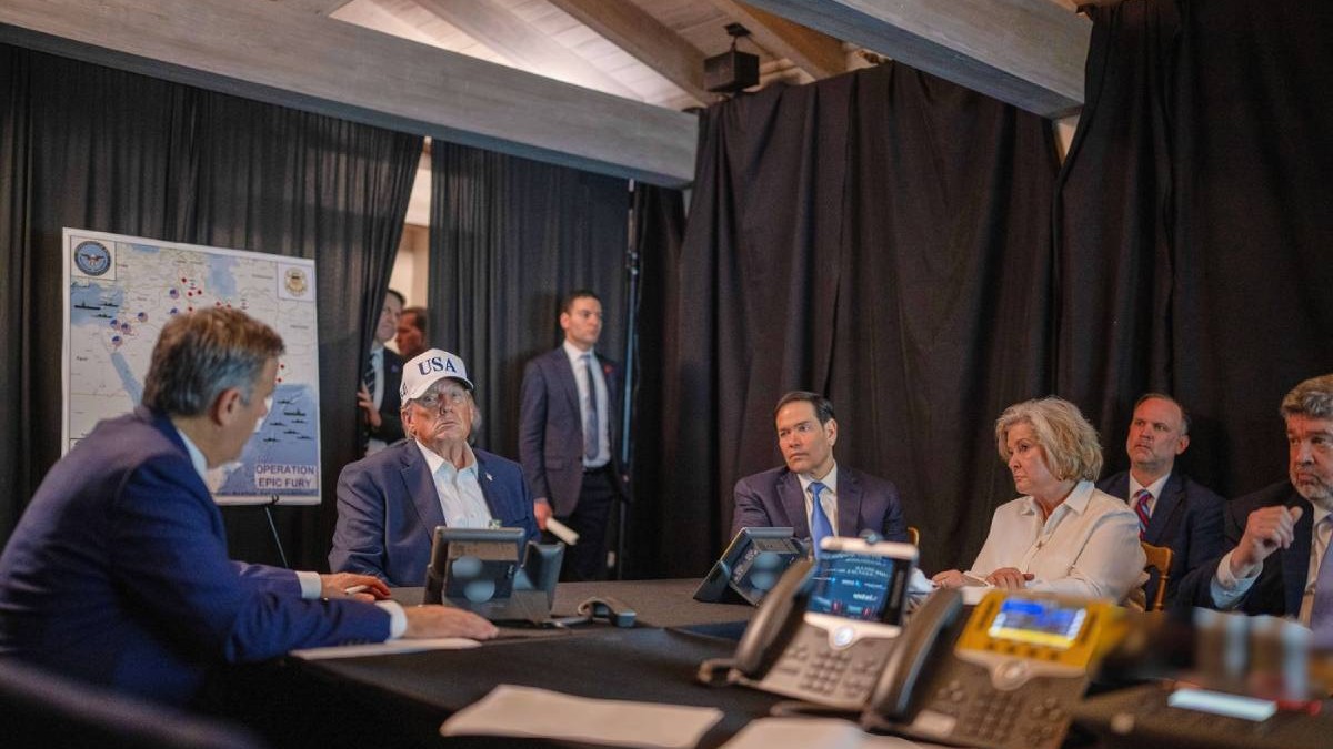 Donald Trump, John Ratcliffe, Marco Rubio, Susie Wiles, se vede o masă lungă de ședință, dosare și sticle de apă pe masă, ecrane și mobilier din lemn în fundal. În imagine apar mai mulți oficiali, iar președintele SUA este surprins în timp ce urmărește operațiunea operațiunea împotriva Iranului, cu o șapcă roșie și cravată, februarie 2026