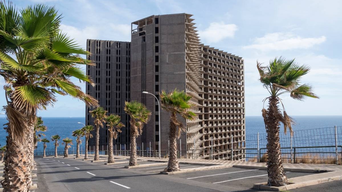 Hotelul Añaza, abandonat și neterminat - Acoran, Tenerife, Spania, se vede constructia, in departare marea, palmieri in prim plan pe marginea unui drum, cerul este senin si este o zi insorita