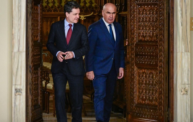 Nicușor Dan și Ilie Bolojan, ambii îmbrăcați în costume elegante, ieșind împreună dintr-o încăpere printr-o ușă masivă din lemn sculptat.