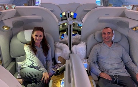 Un bărbat și o femeie la bordul unui avion, stau în fotoliile de la Clasa I și zâmbesc în fața camerei