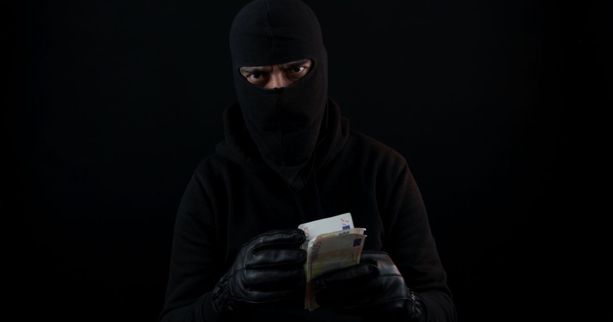 Un bărbat îmbrăcat în negru, cu mănuși și cagulă, ține în mână mai multe banconote de euro și le numără