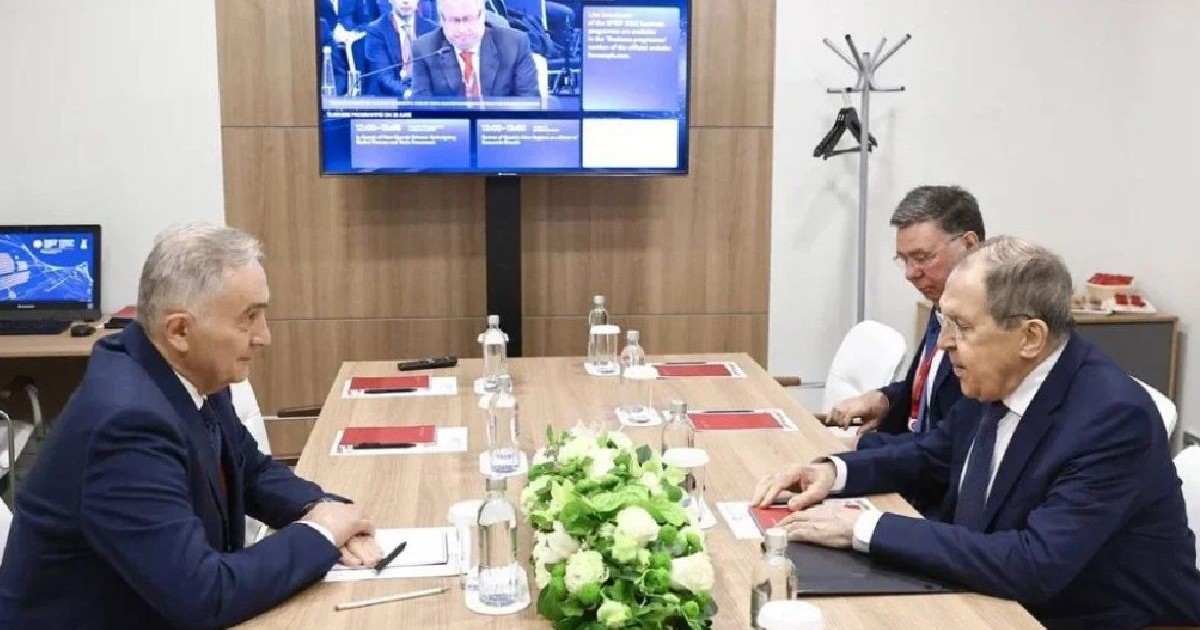 Diplomatul Lazăr Comănescu față în față cu Lavrov la o masa. In plan secundar s emai observa o persoana. Toti cei trei barbati sunt imbracati in costume de culoare inchisa, camasa alba si cravata. In fundal se observa un televizor pe care ruleaza stiri