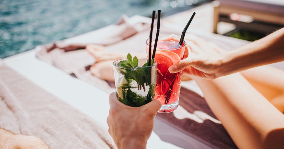 Rețete de cocktail-uri de vară cu rom - Imagine cu un cuplu care ciocneşte pahare cu cocktail pe marginea unei piscine