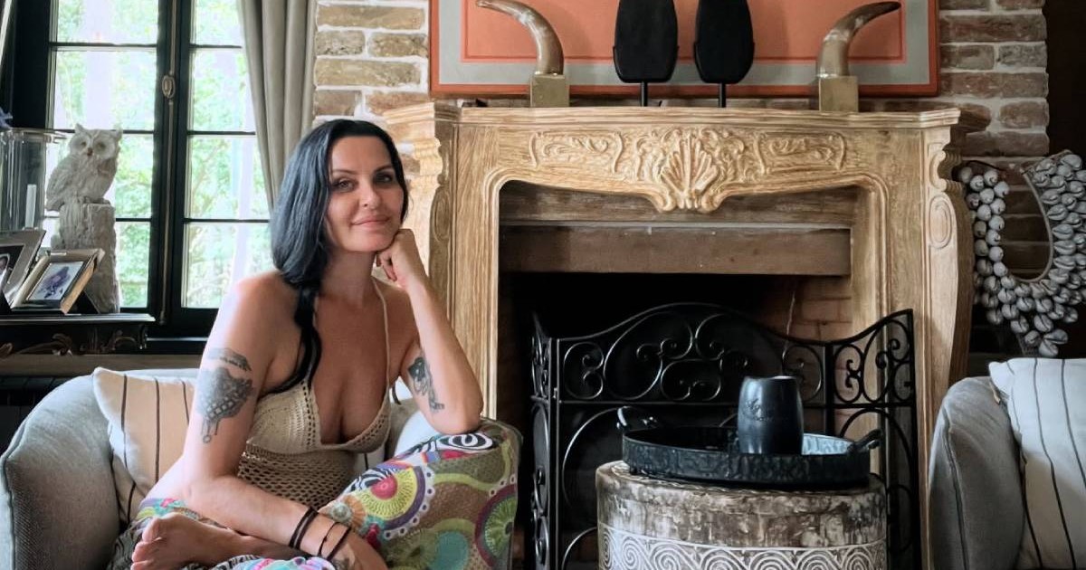 Elena Karaman, cu parul negru si imbracata in tinuta de vara, sta relaxată pe un fotoliu, într-un living elegant, lângă un șemineu decorativ din piatră, într-o casă cu design rustic și luminos.