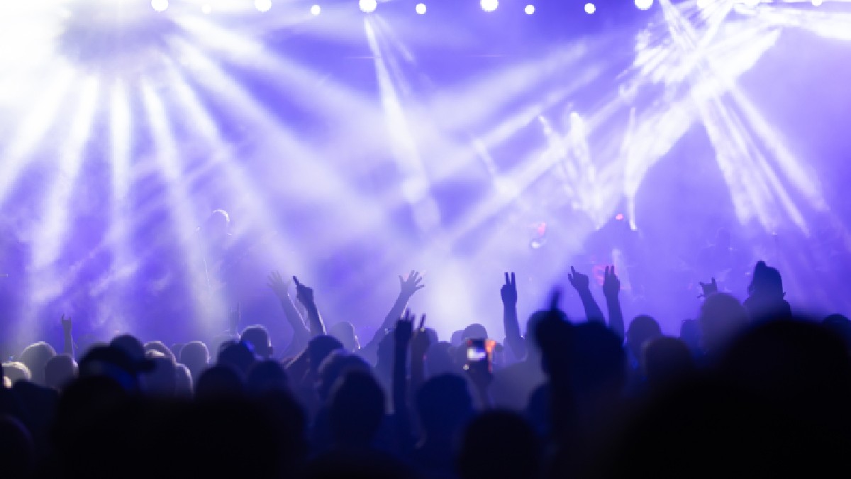 Imaginea surprinde atmosfera energică a unui concert sau a unui eveniment live. În prim-plan se vede o mulțime de oameni, ale căror siluete sunt iluminate de luminile puternice ale scenei. Spectatorii au mâinile ridicate în aer, unii dintre ei făcând un semn de pace, iar altul ține un telefon mobil. Scena este dominată de raze de lumină intense, de culoare albă și albastră, care străpung un strat de fum sau ceață, creând un efect vizual dramatic.