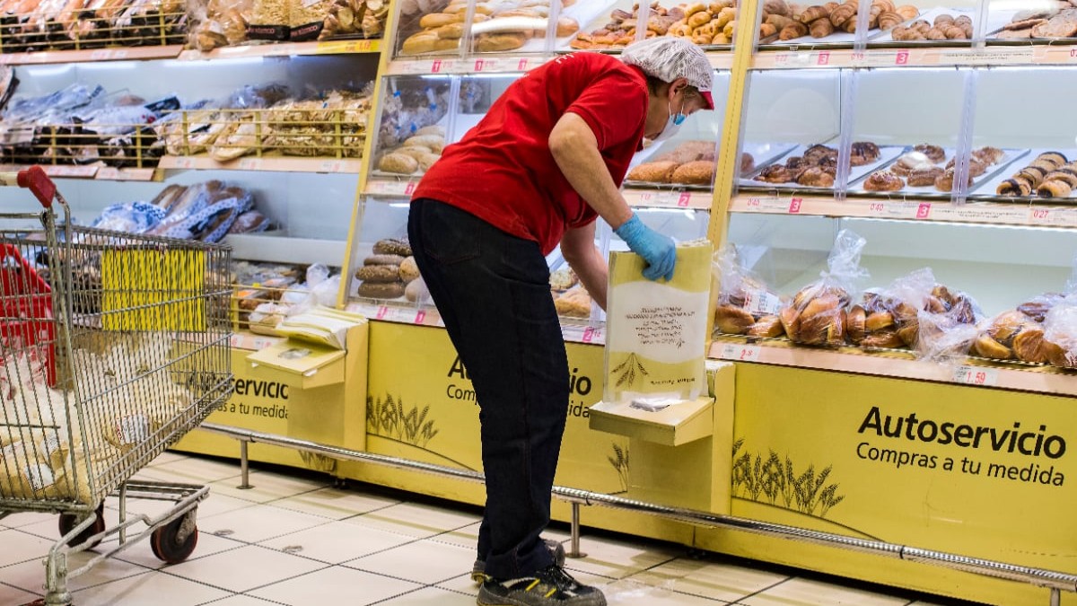 Imaginea prezintă o angajată a unui magazin îmbrăcată în tricou roșu, cu mască și mănuși de protecție, care aranjează pungi în raionul de brutărie cu autoservire.