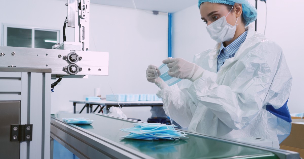 O angajată echipată complet pentru protecție sanitară verifică măști medicale pe o linie automatizată de producție. Imaginea surprinde procesul industrial dintr-o fabrică de echipamente medicale, specific perioadei pandemiei Covid-19.