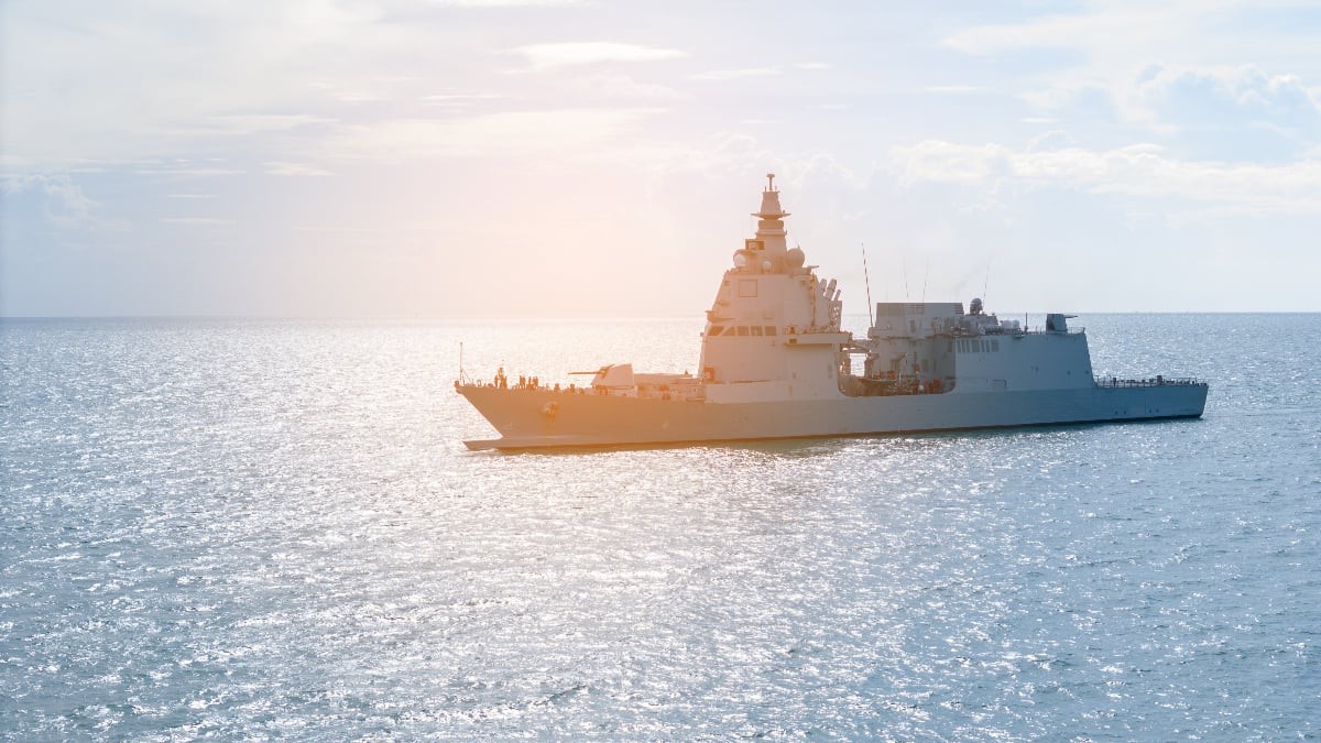 Imaginea surprinde o navă militară de mari dimensiuni navigând pe mare, în condiții de vreme senină, cu soarele reflectându-se pe suprafața apei. Nava este văzută din lateral, fără semne vizibile de avarii, într-un cadru care sugerează o misiune de patrulare sau exercițiu naval.
