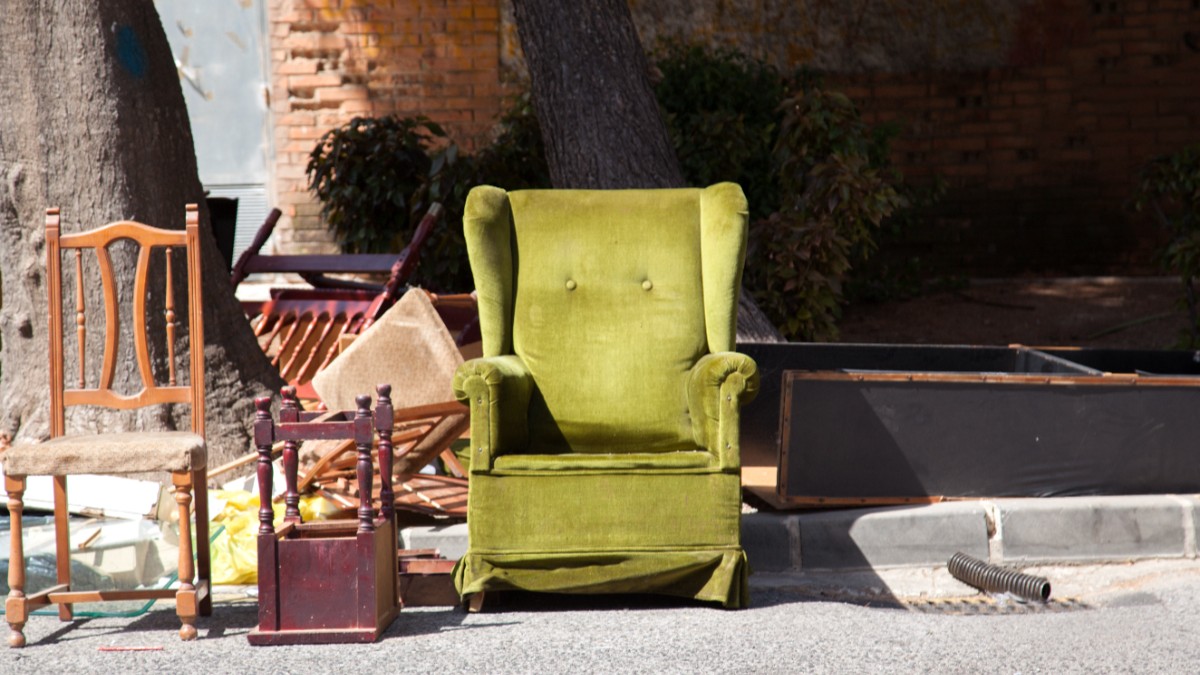 Imaginea prezintă piese de mobilier vechi și abandonate, printre care un fotoliu verde din catifea și un scaun de lemn, lăsate pe marginea trotuarului lângă un zid de cărămidă.