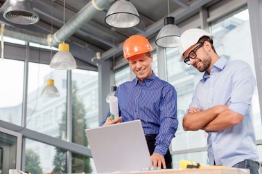 Professional two engineers are discussing the new project. They are using a laptop and smiling. The senior man is standing and holding the blueprint. Copy space in left side