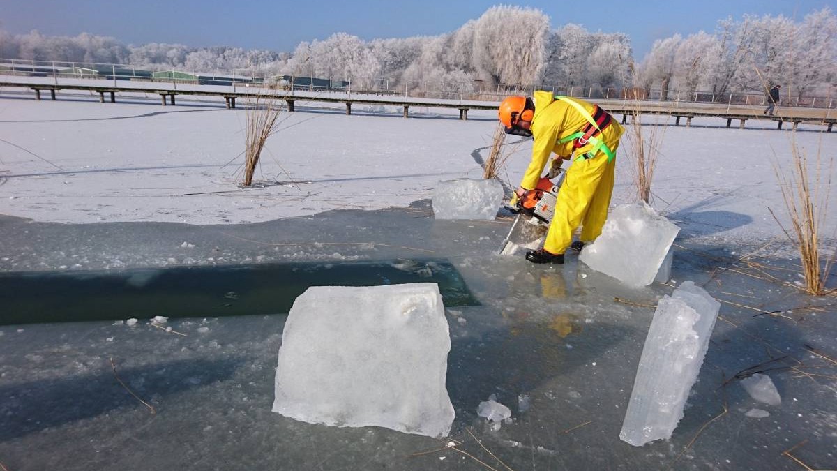 Muncitor de la șantierul de construcții, in costum galben, cu casca potocalie, taie un bloc de gheață cu un ferăstrău cu lanț pe râul Mueritz din Waren, Germania, 14 februarie 2017