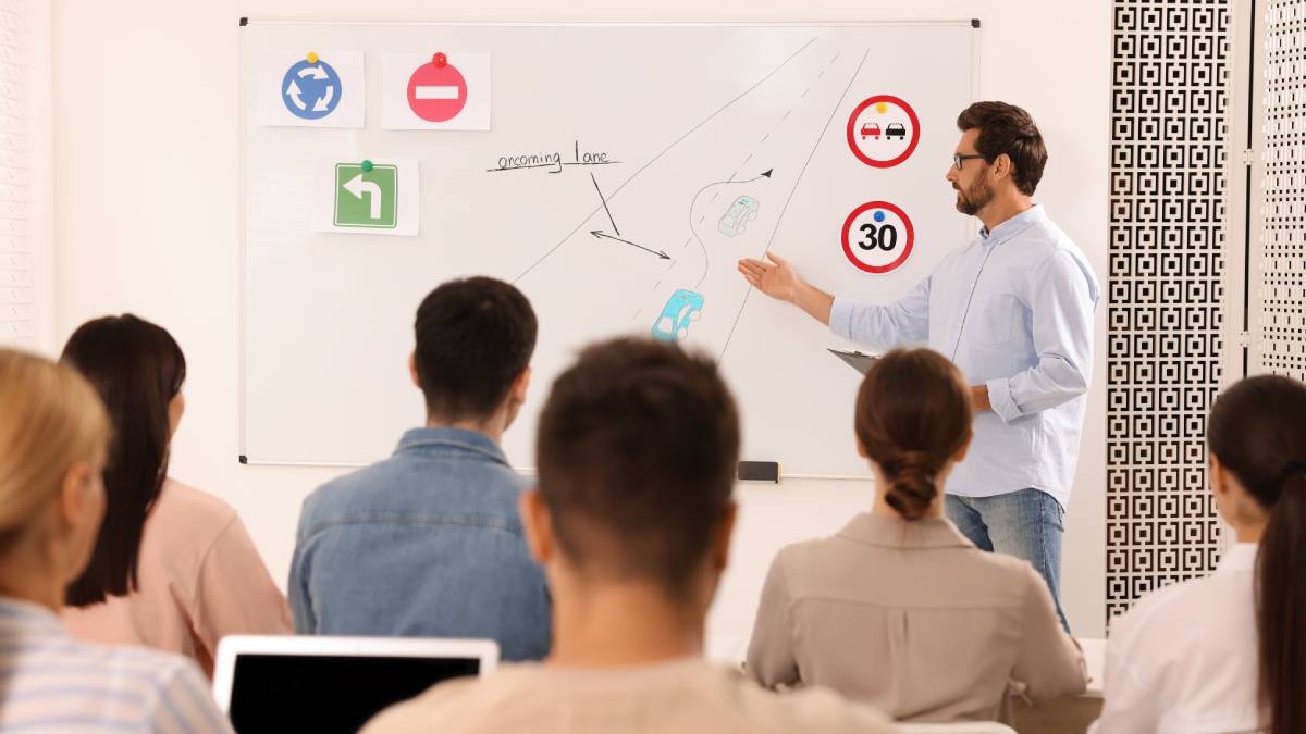 elevi intr-o sala de clasa, in fata lor se afla un profesor care arata spre tabla, unde sunt cateva indicatoare rutiere