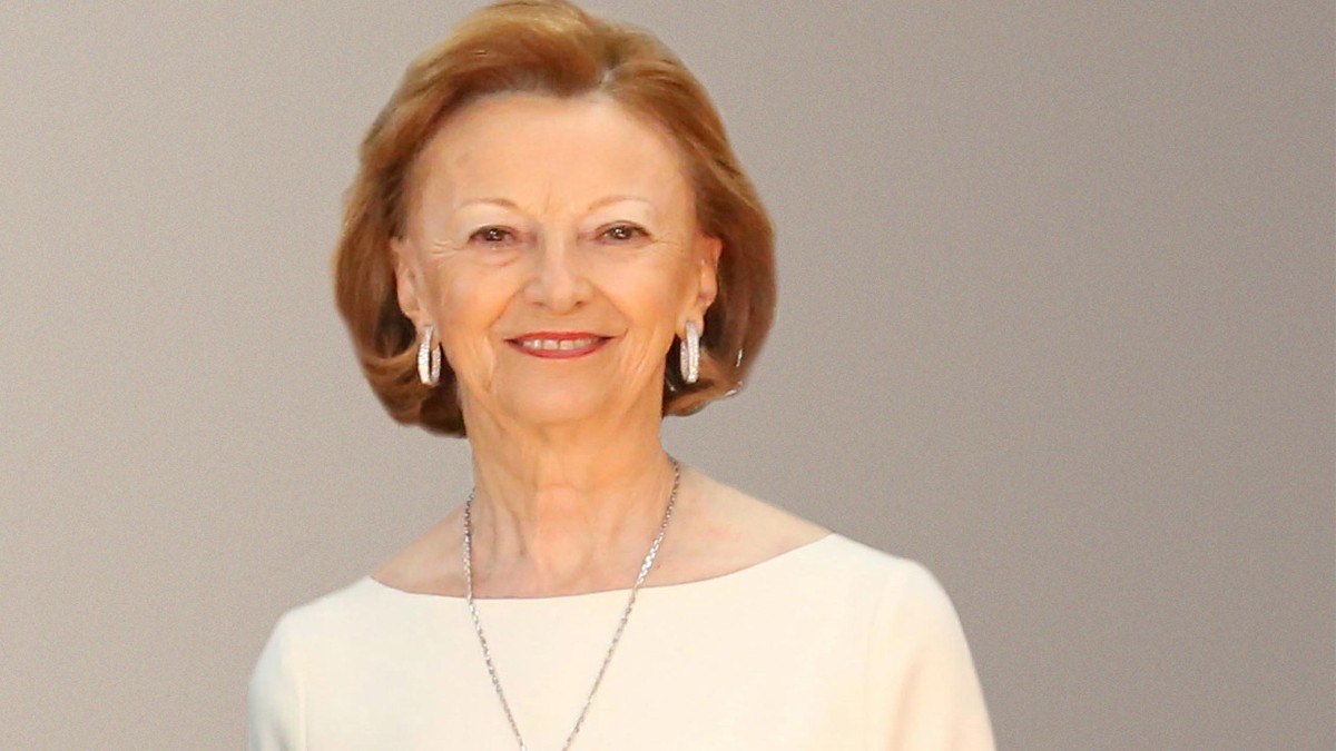 În imagine apare o femeie elegantă, îmbrăcată într-o rochie deschisă la culoare, cu bijuterii discrete și o expresie calmă și zâmbitoare. Portretul sugerează rafinament și sobrietate, fiind realizat într-un cadru simplu, cu fundal neutru.