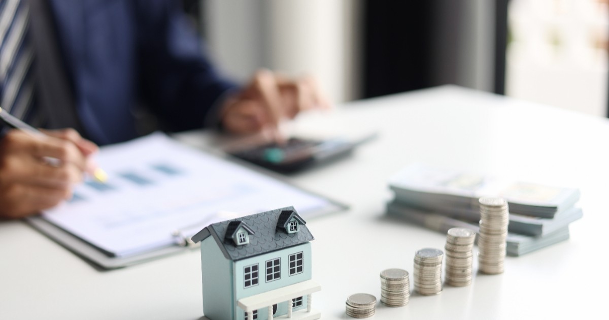 Un agent imobiliar stă la birou cu hârtii în față, are pe masă macheta unei case și mai multe fișicuri de monede așezate unul lângă altul