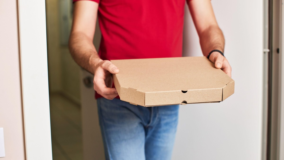 Imagine cu un bărbat, care poartă un tricou roșu și blugi, ținând în mâini o cutie cu pizza