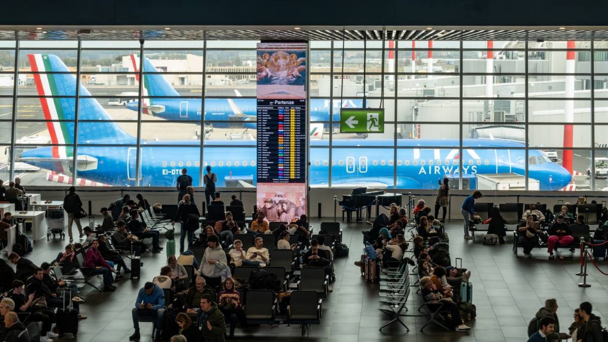 Zeci de persoane care stau și așteaptă la una din porțile de îmbarcarea de pe aeroportul Fiumicino din Italia, în spatele lor se văd avioane vopsite în albastru, parcare și gata să preia pasagerii