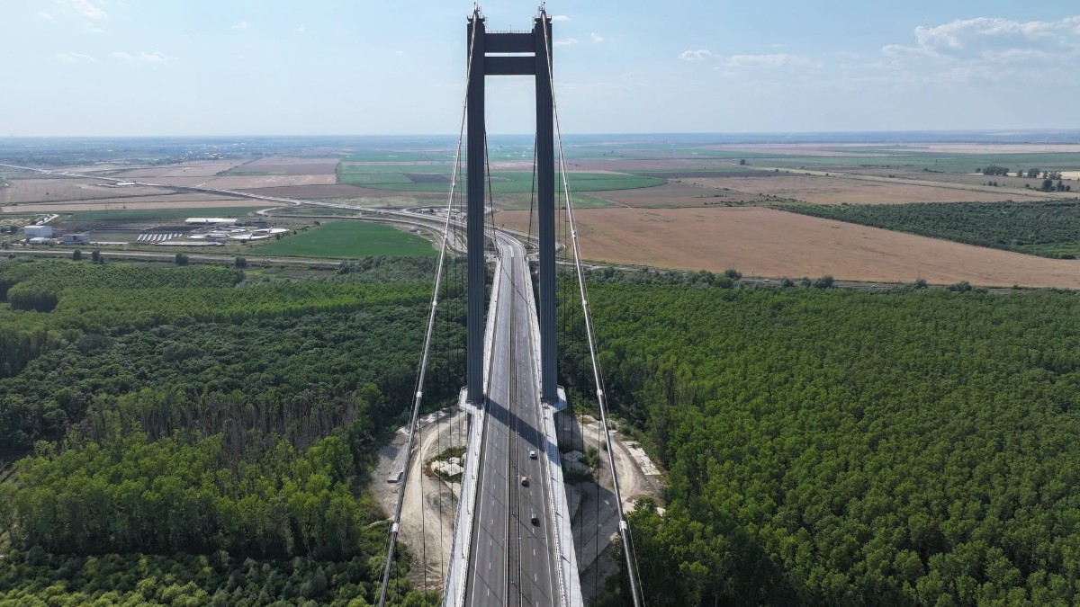 Vedere aeriană a unui pod suspendat care traversează o zonă împădurită bogată și se continuă spre o câmpie întinsă cu terenuri agricole.