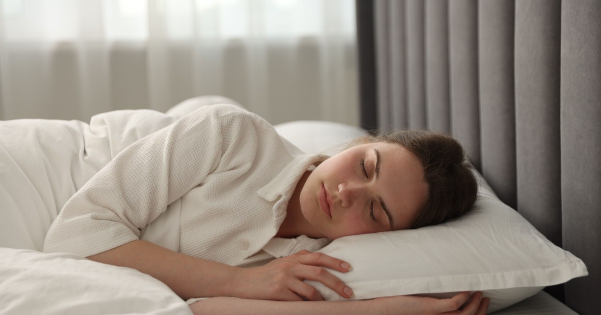 O persoană doarme profund, așezată pe o pernă albă, înfășurată confortabil într-o pilotă albă și purtând un halat alb. Cadul este unul relaxant, cu o tăblie de pat căptușită, de culoare gri închis, și lumina naturală filtrată care pătrunde printr-o fereastră acoperită de draperii deschise în fundal.