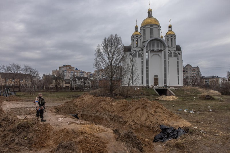 Un membru al armatei ucrainene stă lângă o groapă comună în fața unei biserici ortodoxe, în Bucha, la nord-vest de Kiev (foto EPA)
