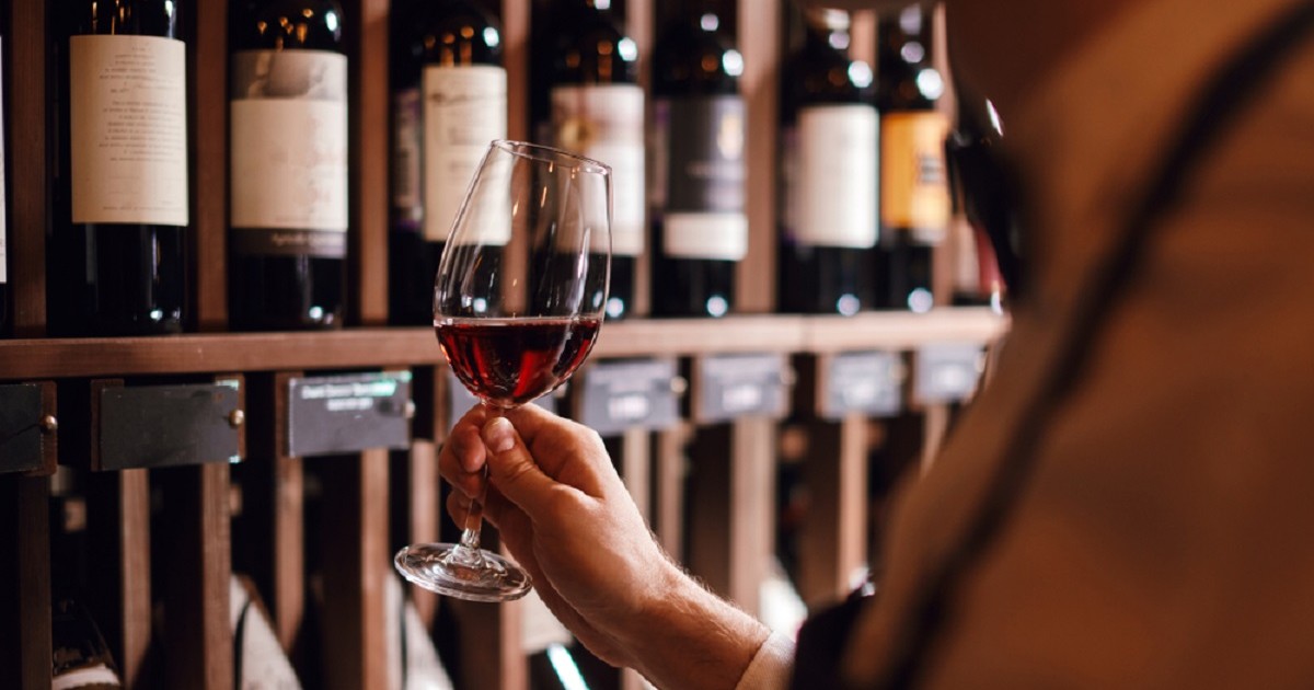 De ce sticla de vin are 750 ml - Un barbat ține în mână un pahar de vin pentru degustare, în fața unor rafturi pline cu sticle de vin