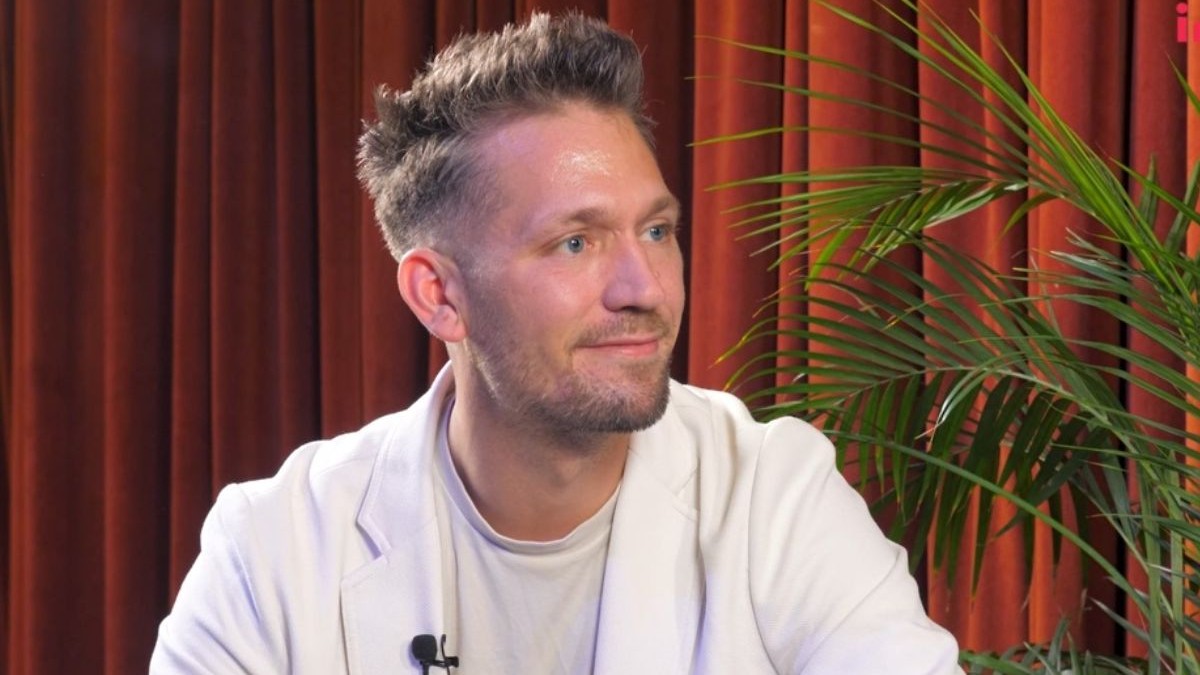Michael Topolinski, îmbrăcat cu un tricou alb și un sacou alb, în timp ce stă pe fotoliu, în platoul Interviurile Imobiliare.ro, cu o perdea roșie pe fundal și o plantă verde