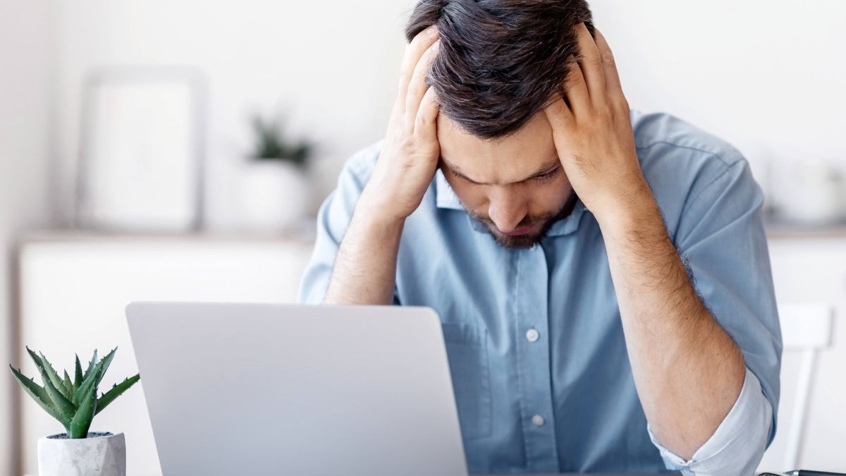 Un bărbat disperat stă la birou în fața unui laptop, în timp ce se ia cu mâinile de cap
