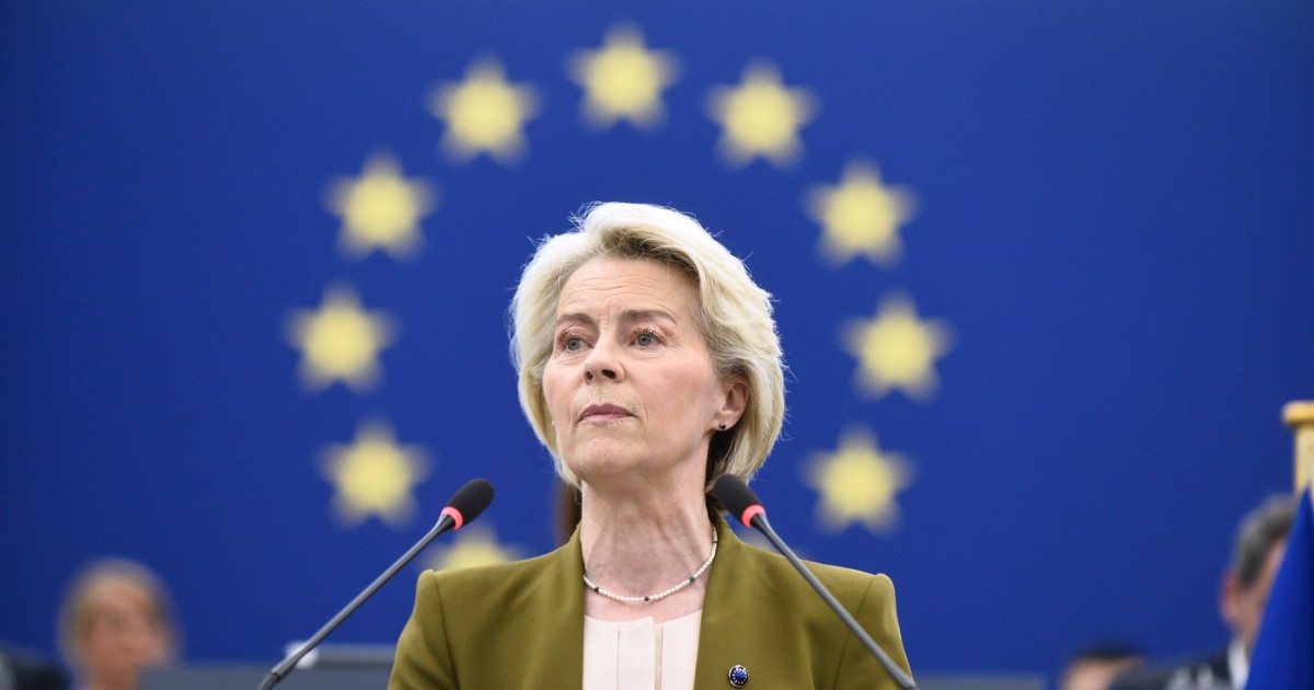 Imaginea o arată pe Ursula von der Leyen vorbind la un pupitru. Are părul scurt, blond și poartă un sacou verde-măsliniu peste un top de culoare crem. Un colier de perle este vizibil la gât. Fundalul este un steag albastru cu un cerc de stele galbene. Două microfoane se află în fața ei Foto: Profimedia