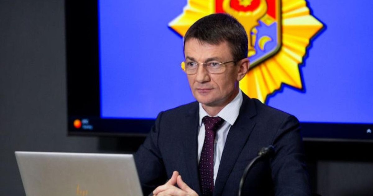 Alecandru Bălan, fost angajat SIS Republica Moldova, îmbrăcat într-un sacou negru, o cămașă albă și o cravată neagră, stând cu mâinile împreunate pe birou, în dreptul pieptului, în fața unui laptop. în spatele său se vede un fundal albastru pe care este proiectată stema Republicii Moldova