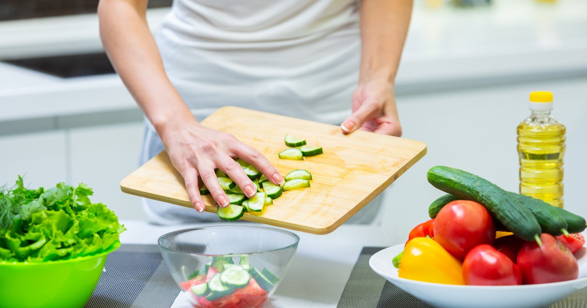 Imaginea surprinde o persoană care pregătește o salată într-o bucătărie modernă, răsturnând felii de castravete de pe un tocător din lemn într-un bol de sticlă. Pe blatul de lucru se mai află un castron verde plin cu frunze de salată verde, un vas alb cu roșii și ardei, și o sticlă de ulei de gătit.