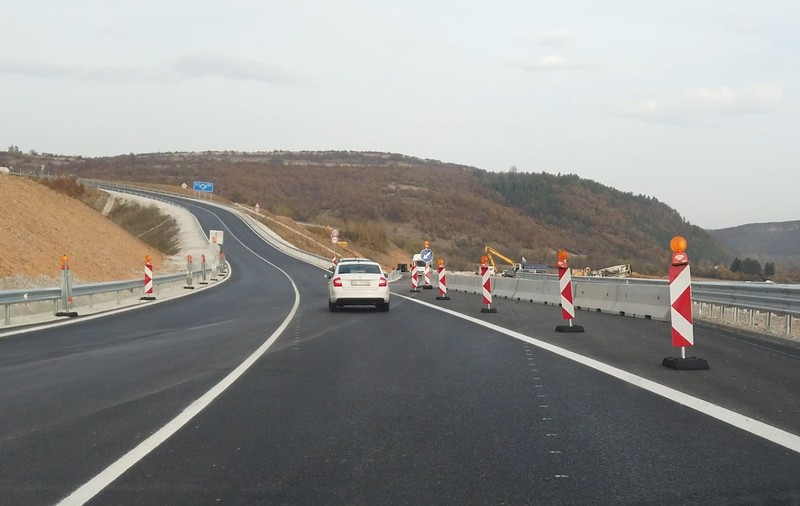 Un autoturism alb care circulă pe o secțiune proaspăt asfaltată de șosea sau autostradă, delimitată de jaloane rutiere și lucrări în desfășurare pe margine.
