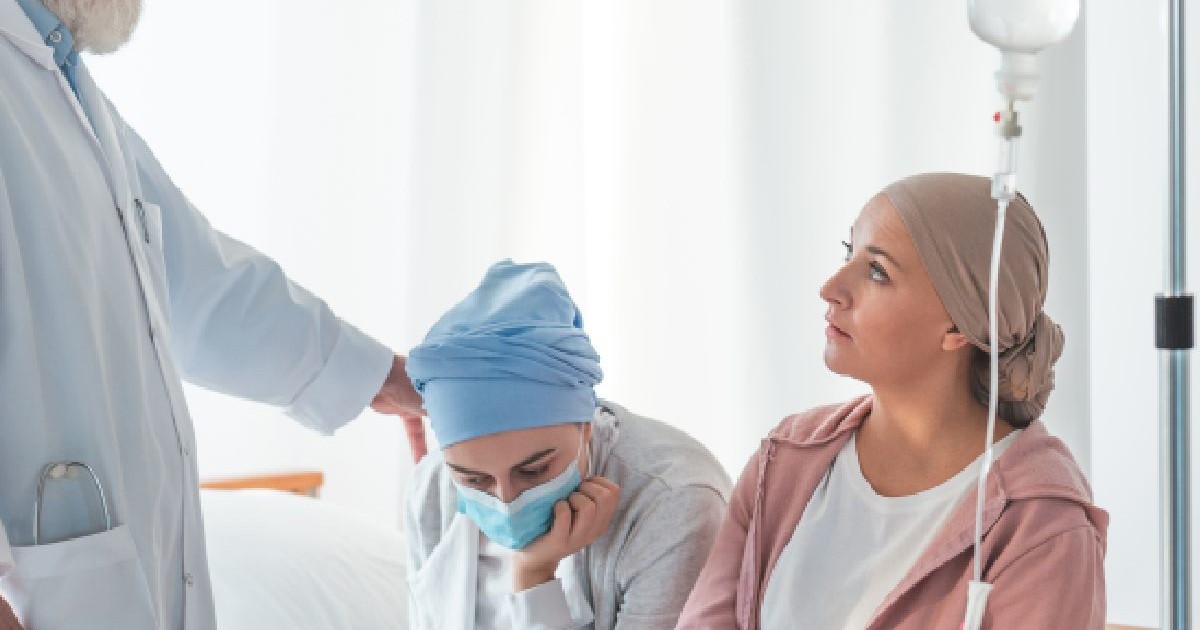Intr-un salon sunt doua femei cu batic pe cap , una dintre ele cu masca, si cu o perfuzie pusa pe stativ, iar o persoana in halat alb ii pune o mana pe umar uneia dintre femei. leucemie transplant medular, centre supraaglomerate