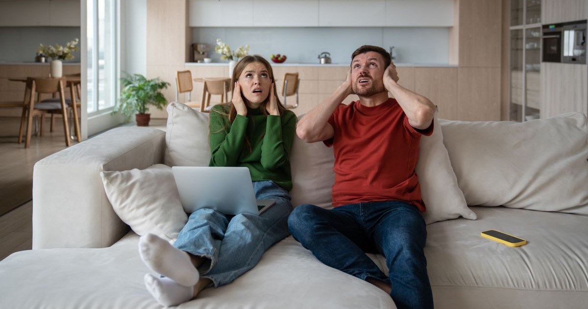 Un cuplu enervat stă pe canapea cu o expresie facială iritată, privind în sus, ascunzându-și urechile din cauza sunetului puternic, a zgomotului din apartamentul vecin.