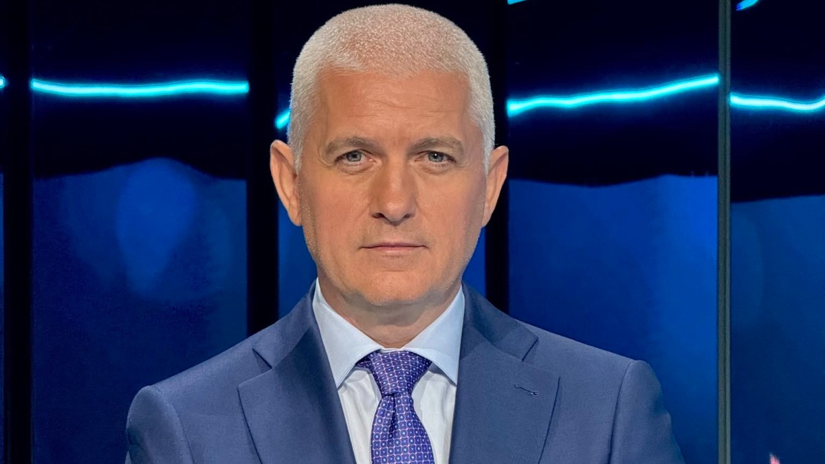 Omul de televiziune Virgil Ianțu este prezentat într-un portret sobru, îmbrăcat într-un costum albastru elegat cu cămașă deschisă și cravată asortată. El privește direct spre cameră cu o expresie serioasă, având ca fundal un studio de televiziune cu lumini albastre.