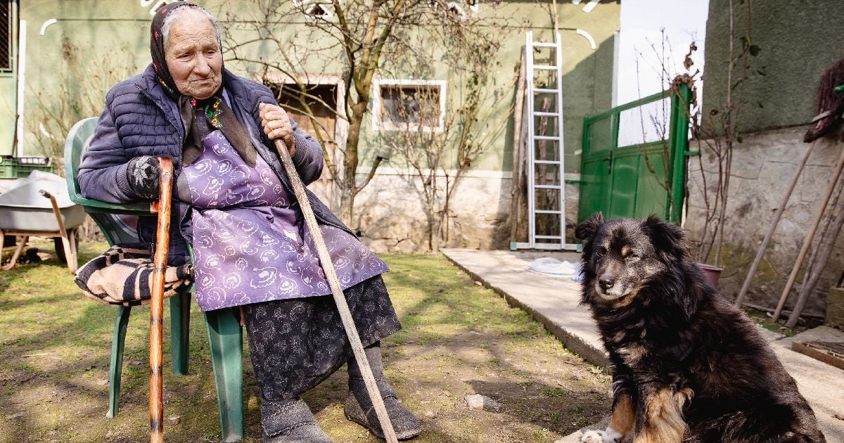 Floarea Bolojan, o femeie în vârstă, mama premierului Ilie Bolojan, stând pe un scaun în curtea casei ei din Birtin, iar alături un câine negru