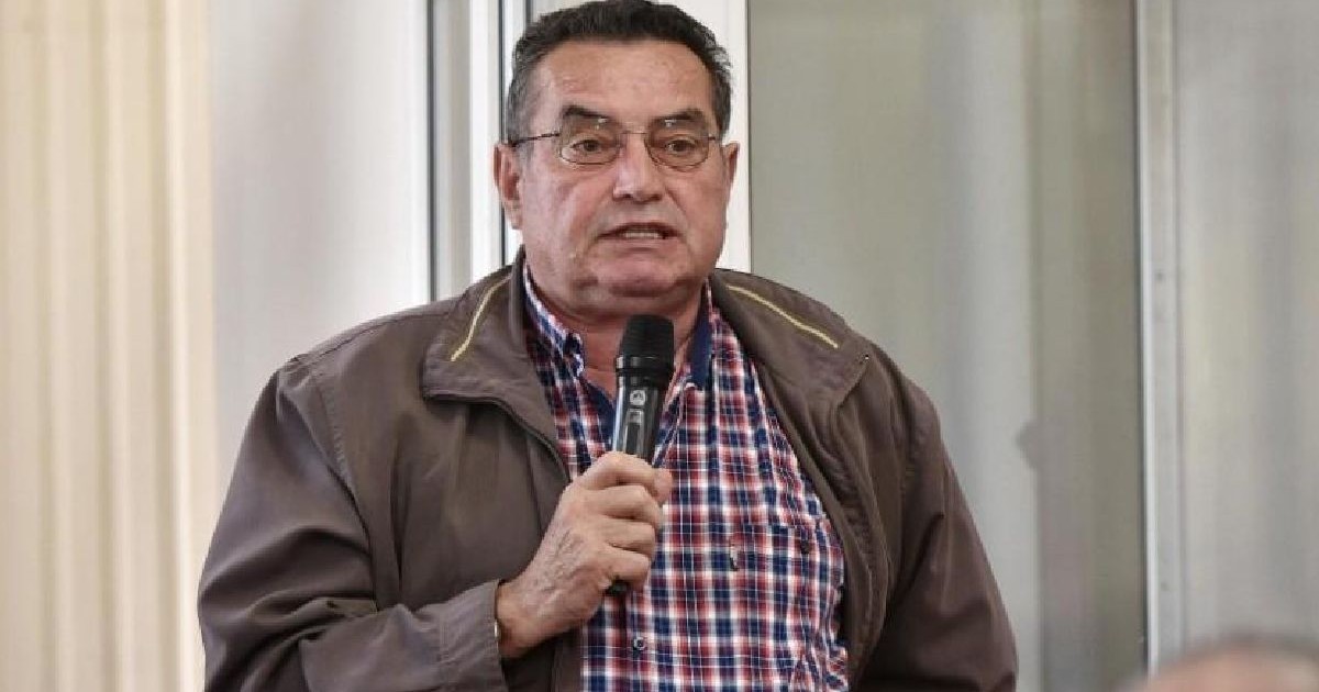 Un bărbat de vârstă mijlocie, cu ochelari, păr scurt și negru și cămașă în carouri, ține un microfon în mână, stând în interior, cu o ușă de sticlă în spatele lui.