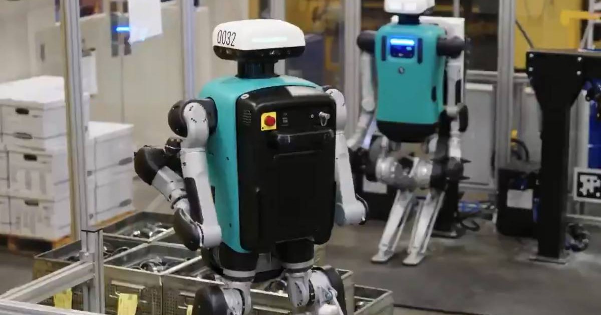 Doi roboți umanoizi Digit, de culoare turcoaz, lucrează într-o hală industrială și mută lăzi din plastic într-o fabrică.