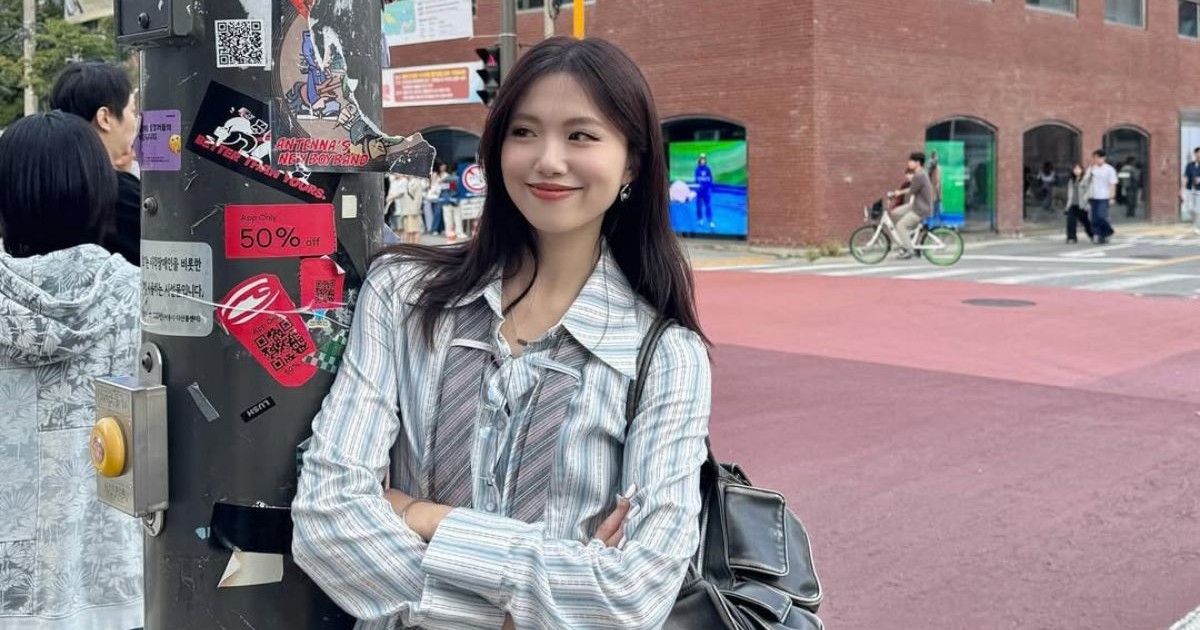 influencerița din Taiwan Irene Gu, purtând o cămașă albă cu dungi gri și o gravată desfăcuta în jurul gâtului, ținând mâinile încrucișate în jurul pieptului și o geantă de piele neagră pe umăr. Femeia stă sprijinită de un stâlp al unui semn de circulație, care este plin de stickere
