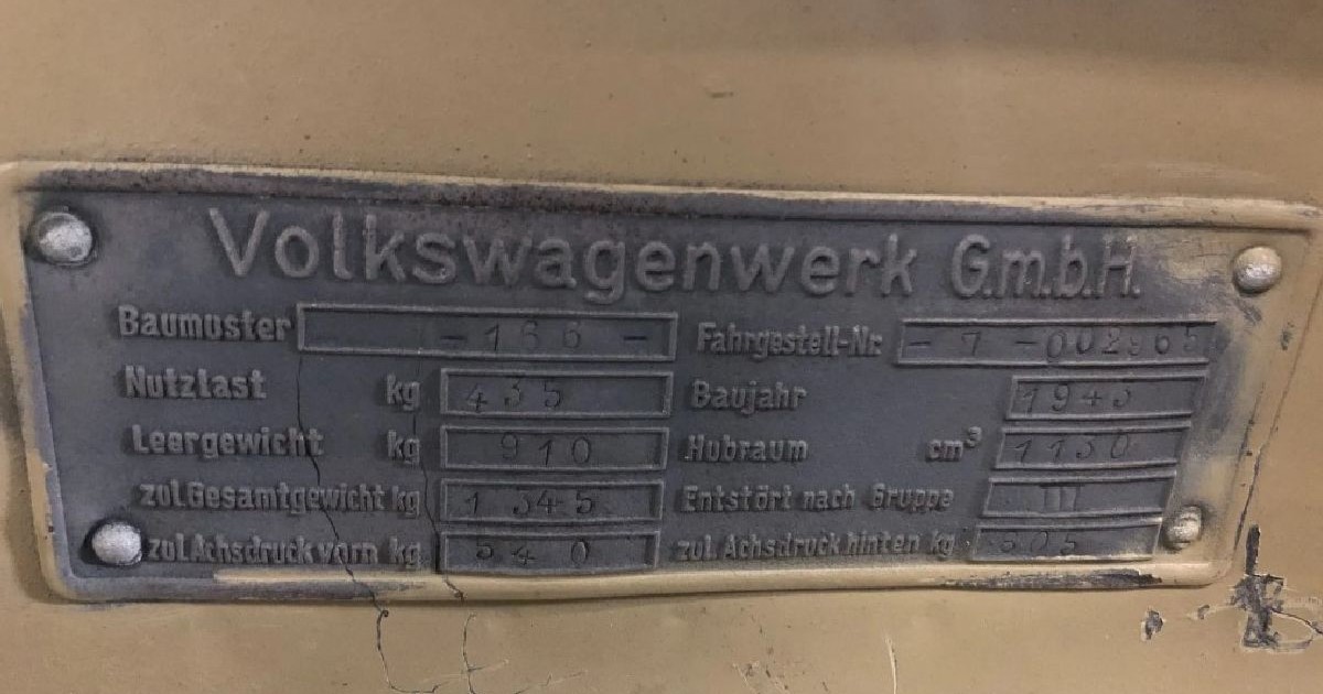 Fotografie a unei plăci metalice vechi de pe motorul unui automobil Volkswagen de epocă, cu specificații tehnice în limba germană, pe care se observă urme de uzură și rugină.